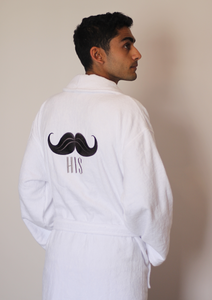 Man wearing white bathrobe with icon of mustache on its back and "his" written under it. He has both his hands in his pockets. Egyptian Cotton bathrobe. 